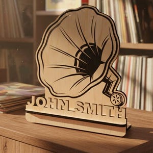 May include: A wooden gramophone-shaped ornament with the name "JOHN SMITH" engraved on the base. The ornament sits on a wooden cabinet alongside a record player and vinyl records. The room has a vintage aesthetic with framed posters and a bookshelf.