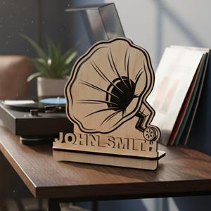 May include: A wooden gramophone-shaped decor piece with the name "JOHN SMITH" engraved on the base. The decor is light brown with black detailing and sits on a dark brown table next to a record player and vinyl records.