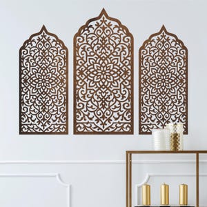 Op de afbeelding: Drie bruine houten wandpanelen met ingewikkelde, opengewerkte ontwerpen. Elk paneel heeft een gewelfde bovenkant en een herhalend bloemmotief. De panelen zijn op een witte muur gemonteerd en creëren een decoratieve en artistieke weergave.