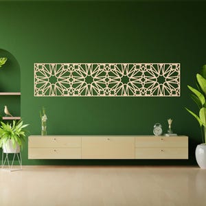 May include: A light-coloured, geometric wooden wall art piece with a repeating star and geometric pattern. The artwork is rectangular and hangs on a dark green wall above a light wood cabinet. The room is decorated with plants and a clock.