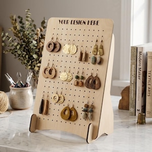 May include: A wooden earring display stand with multiple pairs of earrings. The stand is light-colored with a grid of holes for hanging earrings. The text "YOUR DESIGN HERE" is at the top. Various styles of earrings are displayed, including wood, metal, and stone.