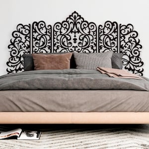 May include: A dark brown, ornate headboard with a scrolling, symmetrical design is mounted above a bed. The bed is made with a light brown base, gray bedding, and decorative pillows in shades of pink and black and white.