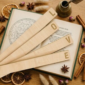 May include: Four light-colored wooden bookmarks arranged in a fan shape, each featuring a single engraved letter spelling out the word LOVE. The bookmarks are resting on an open book with a celestial star chart illustration and printed text.