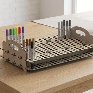 May include: A light-colored wooden marker organizer tray featuring two stacked horizontal panels with numerous circular cutouts for vertical storage. The unit includes side handles and holds a variety of colorful and grey OLO brand markers.