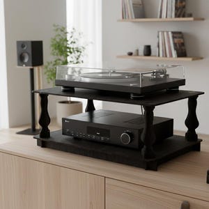 May include: A two-tiered black wooden audio equipment stand featuring four decorative turned legs. The top shelf holds a turntable with a clear dust cover, while the bottom shelf supports a black audio receiver with a front-facing control dial.