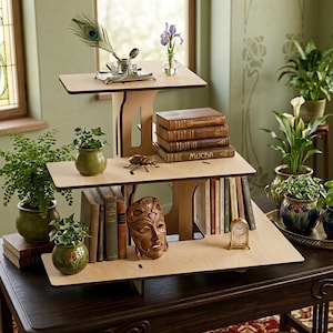 Puede incluir: Una estantería de madera de tres niveles con un acabado natural. Los estantes contienen libros, pequeñas plantas en macetas y objetos decorativos como una máscara y un reloj. Una pluma de pavo real y un tintero se encuentran en el estante superior.