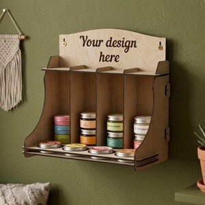 May include: A light-colored wooden display shelf with four vertical compartments and a top panel featuring the text "Your design here". The unit has circular cutouts on the base for holding small round tins, with several colorful tins visible.