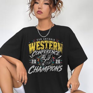 Könnte beinhalten: Ein schwarzes Kurzarm-T-Shirt mit einem Grafikdruck, der SAN ANTONIO WESTERN CONFERENCE CHAMPIONS 20 26 zeigt. Das Design enthält einen Basketball und das Logo der San Antonio Spurs in Gelb, Weiß und Schwarz mit abgenutzten Texturen.
