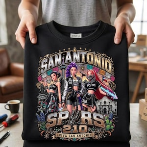 May include: A black crewneck sweatshirt featuring a graphic print of three anime-style figures, the text SAN ANTONIO, SPURS, 210, and VIVA SAN ANTONIO. The design includes illustrations of the Alamo, the Tower of the Americas, and floral accents.