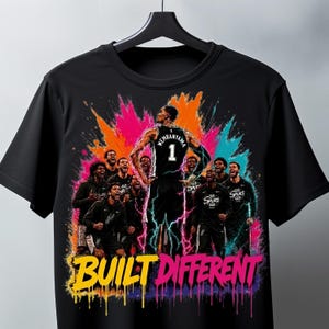 Könnte beinhalten: Ein schwarzes T-Shirt mit einem Grafikdruck eines Basketballspielers mit dem Namen Wembanyama und der Nummer 1 auf dem Trikot, umgeben von einem Team in schwarzen Uniformen, vor einem bunten Farbspritzer-Hintergrund mit dem Text BUILT DIFFERENT.