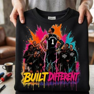 May include: A black crewneck sweatshirt featuring a graphic of basketball player Victor Wembanyama in a Spurs jersey with the number 1, surrounded by teammates, set against a vibrant orange, pink, and blue paint splatter design with the text BUILT DIFFERENT.