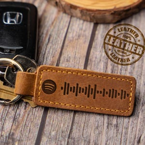 May include: A brown leather keychain with a Spotify code engraved on it. The keychain is rectangular with a loop for keys and a stitched border. The image also shows a car key and other keys.
