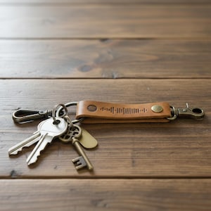 May include: A brown leather keychain with a metal clasp and a ring holding several keys. The leather strap features a Spotify code and a metal button. The keys are silver and bronze, resting on a wooden surface.