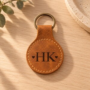 May include: A round, tan leather keychain featuring a dark brown metal ring at the top. The center of the keychain is embossed with the letters HK, flanked by two small heart symbols, and finished with a light-colored stitched border.