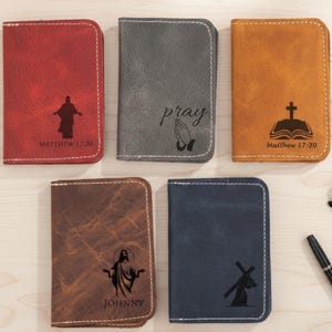 May include: A collection of five leather wallets in various colors: red, gray, tan, brown, and navy blue. Each wallet features a unique religious design, including images of Jesus, praying hands, a cross, and text from Matthew 17:20 and the name "Johnny".