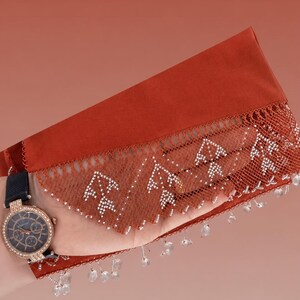 May include: A rust-coloured textile with a delicate lace trim, adorned with small white beads and dangling clear crystal accents. A gold-toned watch with a black band is visible on the wrist. The watch face has a dark blue background and is encrusted with small crystals.