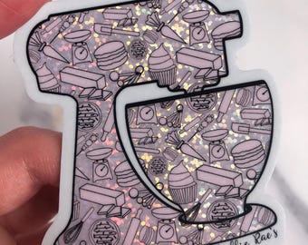 May include: A sticker in the shape of a stand mixer, featuring a pattern of baking tools and treats in pink and silver. The design includes cupcakes, scales, and rolling pins. The text "Mollie Rae's" is printed on the sticker.