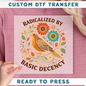 a hand holding a transparent square with a colorful illustration of a bird surrounded by flowers and leaves.