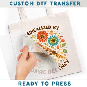 A hand is holding a white tote bag with a colorful floral design and the text "RADICALIZED BY BASIC DECENCY" printed on it.