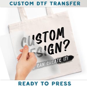 A hand is holding a piece of paper with the text "CUSTOM DTF TRANSFER" printed on it, and the person is pressing it against a white background.