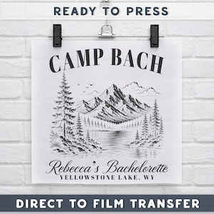 May include: A white square direct to film transfer sheet featuring a black ink illustration of a mountain range, pine trees, and a lake. The text reads CAMP BACH, Rebecca's Bachelorette, and YELLOWSTONE LAKE, WY. The sheet is held by a black hanger.