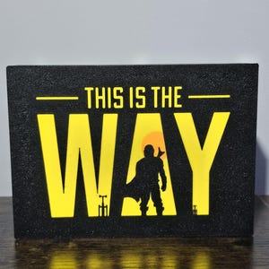 Könnte beinhalten: Schwarzes rechteckiges Schild mit gelbem Text, der "THIS IS THE WAY" lautet. Das Wort "WAY" steht in großen, fetten Buchstaben, mit einer Silhouette einer Figur in der Mitte. Der Hintergrund hinter der Figur ist ein gelber Kreis.