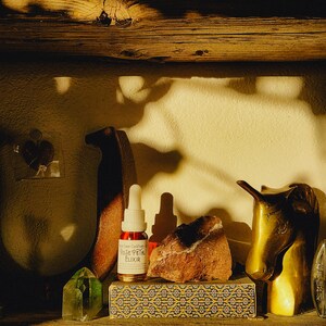 May include: A wooden shelf displays a Rose Petal Elixir bottle, a green crystal, a decorative box, a rock, and a brass horse head. The elixir bottle has a dropper and a label. The shelf is made of wood.