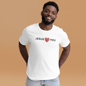 May include: A white short-sleeved t-shirt featuring the black text JESUS and YOU with a red geometric heart graphic in the center. The shirt has a crew neckline and a standard fit, displaying a clean design on a plain white fabric base.