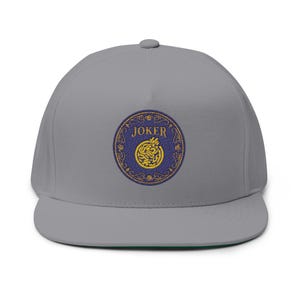 May include: A gray flat-brimmed hat with a circular embroidered patch. The patch features the word "JOKER" in gold lettering around a blue circle with a gold design and a green underside.