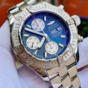 May include: A silver-tone stainless steel Breitling Superocean automatic chronograph watch with a blue dial, three white sub-dials, and a day-date window. The watch features a rotating bezel with engraved numerals and a metal link bracelet.