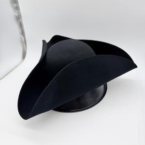 May include: A black tricorn hat, a three-cornered hat, sits on a black stand against a white background. The hat has a rounded crown and three upturned brims. The hat appears to be made of felt or a similar material.