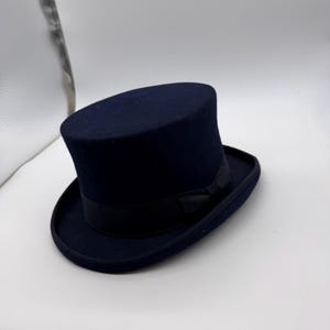 Puede incluir: Un sombrero de copa azul marino con una cinta negra. El sombrero tiene una copa alta y cilíndrica y un ala ancha. Este sombrero formal se usa a menudo para ocasiones especiales.