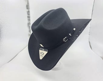 May include: A black cowboy hat with a curved brim and a tall crown. The hat features a decorative band with a silver buckle and accents. A small tag with a logo hangs from the hat. The hat is displayed on a white surface.