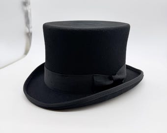 May include: A black top hat with a flat crown and a curved brim. The hat features a matching black grosgrain ribbon band wrapped around the base of the crown, finished with a small, simple bow detail on the side of the hat.