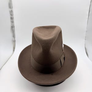 Puede incluir: Un sombrero fedora marrón con una banda marrón oscuro y una pluma. El sombrero tiene una corona clásica en forma de lágrima y un ala ancha. Un accesorio elegante.