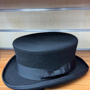 May include: A classic black top hat, featuring a cylindrical crown and a wide brim, adorned with a black grosgrain ribbon. The hat is displayed on a wooden shelf with other hats.