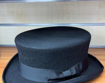May include: A classic black top hat, featuring a cylindrical crown and a wide brim, adorned with a black grosgrain ribbon. The hat is displayed on a wooden shelf with other hats.