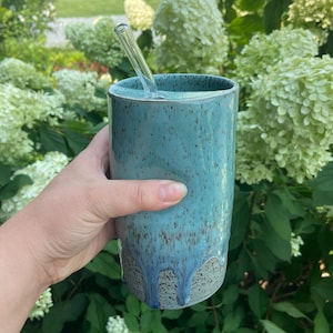 May include: A cylindrical ceramic tumbler with a speckled teal and blue glaze featuring a drip pattern near the base. The tumbler has a matching ceramic lid with a small opening and a clear glass straw inserted into the top for drinking.