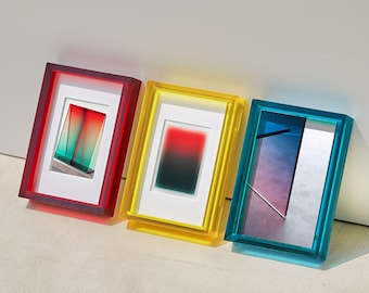 Puede incluir: Tres marcos de arte con marcos de acrílico coloridos en rojo, amarillo y azul. Cada marco contiene una imagen abstracta única con un passe-partout blanco. Los marcos están apoyados contra una pared blanca.