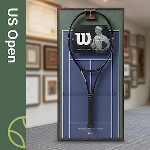 Puede incluir: Una exhibición enmarcada de una raqueta de tenis negra Wilson centrada sobre un diseño de cancha de tenis azul. La exhibición incluye una foto de un tenista y el texto "US Open". El marco es marrón oscuro.