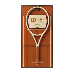 Puede incluir: Una raqueta de tenis con marco blanco y empuñadura marrón se muestra sobre un fondo marrón, que se asemeja a una cancha de tenis. La raqueta tiene el logotipo "Wilson" y la marca "Roland Garros". El texto "CLASH" también es visible.