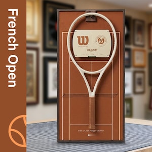 Puede incluir: Una raqueta de tenis enmarcada con marco blanco y empuñadura marrón, exhibida sobre un fondo marrón que representa una cancha de tenis. El marco incluye el texto "French Open" y los logotipos de Wilson y Roland Garros.