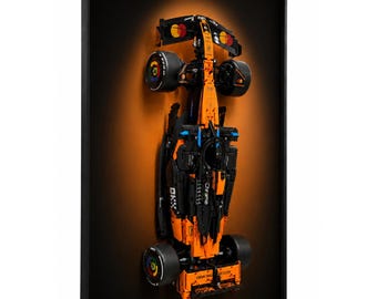 May include: A framed image of a Formula 1 race car model. The model is primarily orange and black, with visible details like wheels and sponsor logos. The background is a gradient of orange and black.