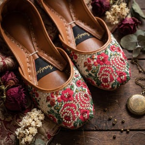 May include: A pair of slip-on flats featuring a cream-colored fabric upper adorned with intricate red floral embroidery, green leaves, and small clear beads. The interior is lined with tan leather and includes a black heel patch with white text.