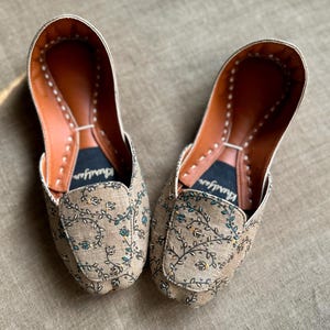 May include: A pair of tan slip-on shoes with floral embroidery. The shoes have a brown leather interior and a stitched trim. The floral pattern includes green and blue accents. The brand name "Khairifar" is printed on the insole.