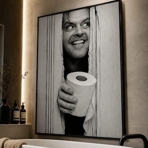 May include: A black and white framed wall art print featuring a man with a wide, intense expression peering through a vertical gap in a wooden door, holding a roll of toilet paper in the foreground. The artwork is displayed in a thin black frame.