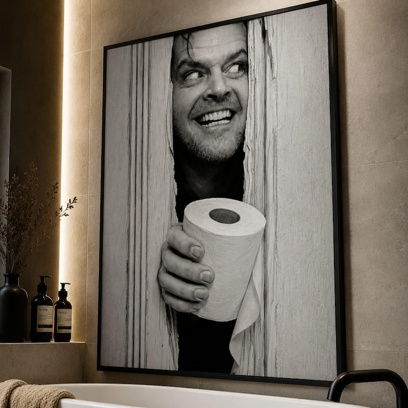 May include: A black and white framed wall art print featuring a man with a wide, intense expression peering through a vertical gap in a wooden door, holding a roll of toilet paper in the foreground. The artwork is displayed in a thin black frame.