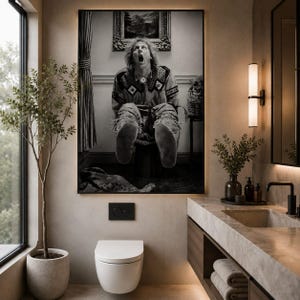 May include: A black and white wall art print featuring a person with long hair and a patterned shirt sitting on a toilet with their mouth open in a shocked expression. The artwork is framed and displayed above a modern white wall-mounted toilet.