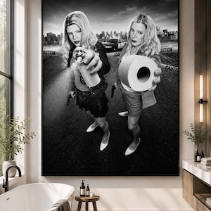 May include: A black and white wall art print depicting two identical blonde figures standing on asphalt. One figure holds a spray paint can, while the other holds a roll of toilet paper. A police car and caution tape are visible in the background.