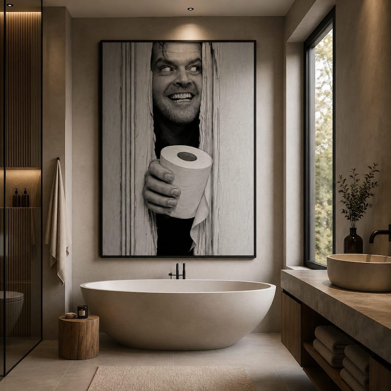 May include: A large black and white wall art print featuring a man peering through a broken door frame while holding a roll of toilet paper. The artwork is displayed above a modern oval-shaped bathtub in a neutral-toned bathroom setting.